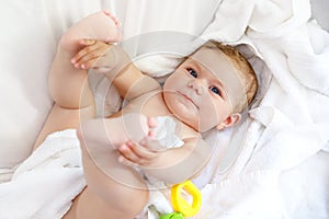 Cute little baby playing with toy rattle and own feet after taking bath. Adorable beautiful girl wrapped in white towels