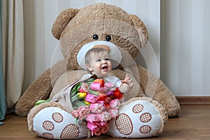 Cute little baby smiling with his first teeth, sitting on huge teddy bear with big bouquet of tulips