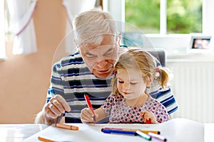 Cute little baby toddler girl and handsome senior grandfather painting with colorful pencils at home. Grandchild and man