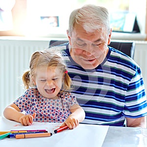 Cute little baby toddler girl and handsome senior grandfather painting with colorful pencils at home. Grandchild and man