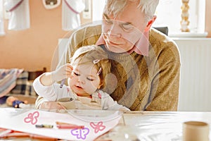 Cute little baby toddler girl and handsome senior grandfather painting with colorful pencils at home. Grandchild and man