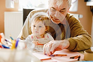 Cute little baby toddler girl and handsome senior grandfather painting with colorful pencils at home. Grandchild and man