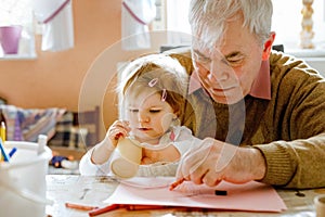 Cute little baby toddler girl and handsome senior grandfather painting with colorful pencils at home. Grandchild and man