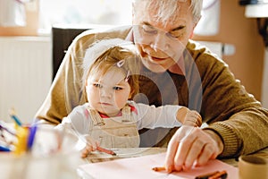 Cute little baby toddler girl and handsome senior grandfather painting with colorful pencils at home. Grandchild and man having