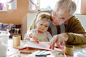 Cute little baby toddler girl and handsome senior grandfather painting with colorful pencils at home. Grandchild and man