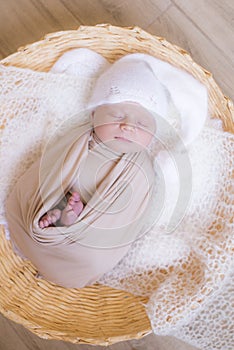 Cute little baby in white knitted hat lies in a wicker basket in a beige knitted blanket. Summer mood.