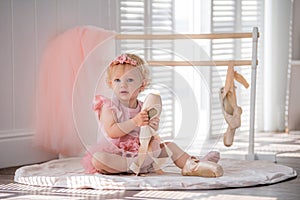 A cute little ballerina in a pink ballet costume sits near the barre in the room and tries to put on her pointe shoes. Kid and