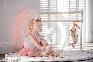 A cute little ballerina in a pink ballet costume sits near the barre in the room and tries to put on her pointe shoes. Kid and