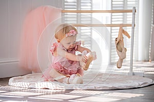 A cute little ballerina in a pink ballet costume sits near the barre in the room and tries to put on her pointe shoes. Kid and
