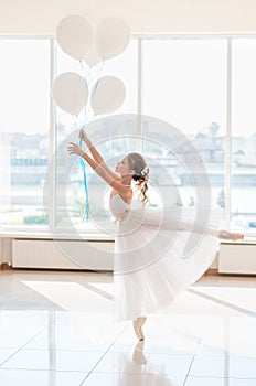 Cute little ballerina in white dress and pointe shoes with white balloons is dancing in the room