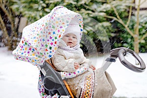 Cute little beautiful baby girl sitting in the pram or stroller on cold snowy winter day. Happy smiling child in warm