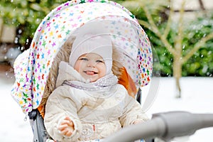 Cute little beautiful baby girl sitting in the pram or stroller on cold snowy winter day. Happy smiling child in warm