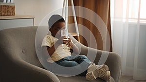 Cute Little Black Boy Drinking Water From Glass While Relaxing At Home