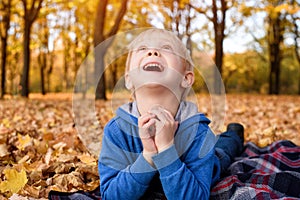 Cute little blond boy lies on a plaid and look up, yellow autumn leaves. Smiling and having fun. Fall day