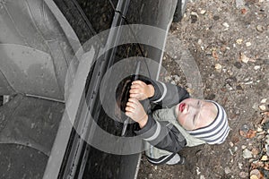 Cute little blond boy in striped hat struggles to open door of black, dusty car, hanging on handle. Family travel