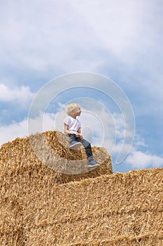 Cute little blond boy in white T-shirt is sitting on haystack on field. Boy looking into the distance. Summer holidays
