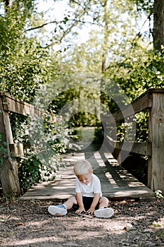 Cute Little Blond Haired Toddler Boy Kid Child Sitting and Laughing in Front of Wooden Bridge Over a Creek at the Outdoor Park in