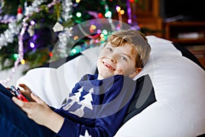Cute little blond kid boy playing with a video game on gadget console on Christmas with decorated tree on background