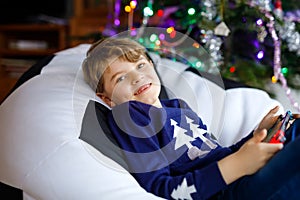 Cute little blond kid boy playing with a video game on gadget console on Christmas with decorated tree on background