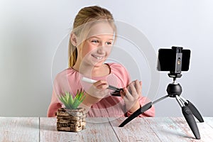 Cute little blonde beauty blogger. Girl speaking in front of the camera for Vlog. A beautiful smiling schoolgirl sits at a table