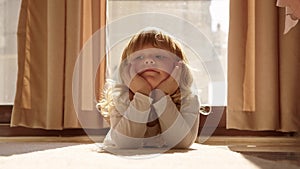 cute little blonde girl lie on carpet at home on floor and props her face with her hands, looks at camera with tired but