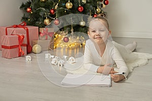 Cute little blonde girl lying on the floor near the Christmas tree writing a letter to Santa Claus.