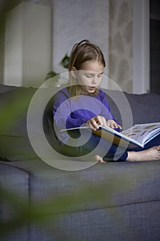 A cute little blonde girl is reading a large colorful book sitting on the sofa in the living room. Self-education of