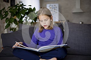 A cute little blonde girl is reading a large colorful book sitting on the sofa in the living room. Self-education of