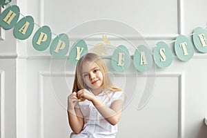 Cute little blonde girl sitting in a bright room near the window on the background of streamers with the words Happy
