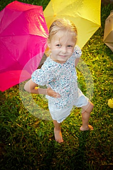 Cute little blonde girl with umbrella under rain drops on lawn in a sunny summer day