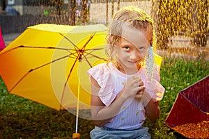 Cute little blonde girl with umbrella under rain drops on lawn in a sunny summer day