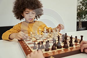 Cute little boy with afro hair thinking before making a move while playing chess, sitting at the table indoors