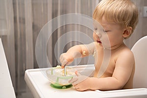 Cute little boy alone eating porridge with a spoon from a plate, healthy eating concept. The kid learns to eat with a spoon on his