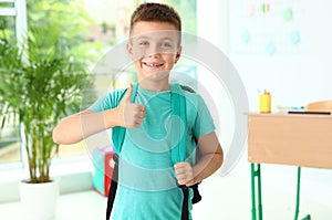 Cute little boy with backpack showing thumbs-up in classroom