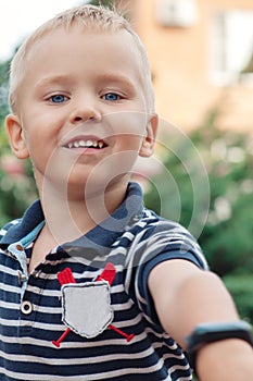 Cute little boy with blond hair and blue eyes reaches out the hand with electronic watch towards the camera, smiling.