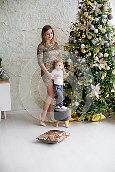 Cute little boy with blond hair plays with his mother in golden shiny dress in a bright room near the Christmas tree