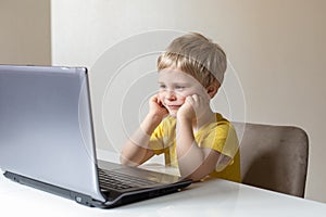 A cute little boy with blond hair sits at a table with a laptop. The boy put his hands on his face and thinks