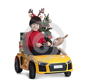 Cute little boy with Christmas tree, deer toy and gift boxes driving children`s car on white background