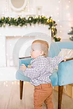 Cute little boy by the Christmas tree and fireplace decorated with garlands and gifts.