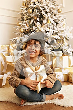 Cute little boy with dark hair in cozy clothes posing near decorated Christmas tree with presents