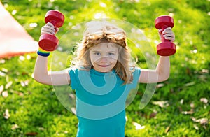 Cute little boy doing exercises with dumbbells in green park. Portrait of sporty child with dumbbells. Happy child boy