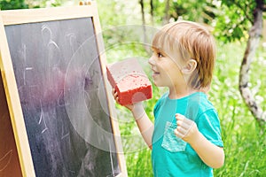 Cute little boy drawing on blackboard with chalk, outdoor on summer sunny day. Back to school concept.