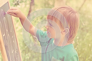 Cute little boy drawing on blackboard with chalk, outdoor on summer sunny day. Toned.