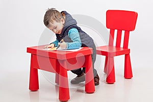 Cute little boy drawing, red table and chair, on white background. Childhood education concept