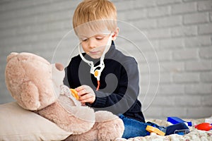 Cute little boy dressed as doctor playing with toy bear at home.