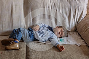 Cute and little boy dressed in blue and brown shoes, is sleeping peacefully on sofa in the living room of his house