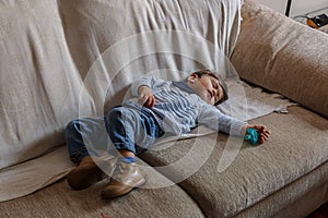 Cute and little boy dressed in blue and brown shoes, is sleeping peacefully on sofa in the living room of his house