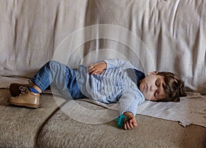 Cute and little boy dressed in blue and brown shoes, is sleeping peacefully on sofa in the living room of his house