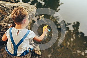 Cute little boy dressed in blue shorts, launch paper ship on the lake in the park. Adorable kid boy playing with a boat. Child