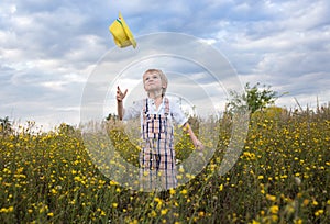 Cute little boy dressed in rustic style tosses his yellow hat while standing in a field of wild yellow flowers